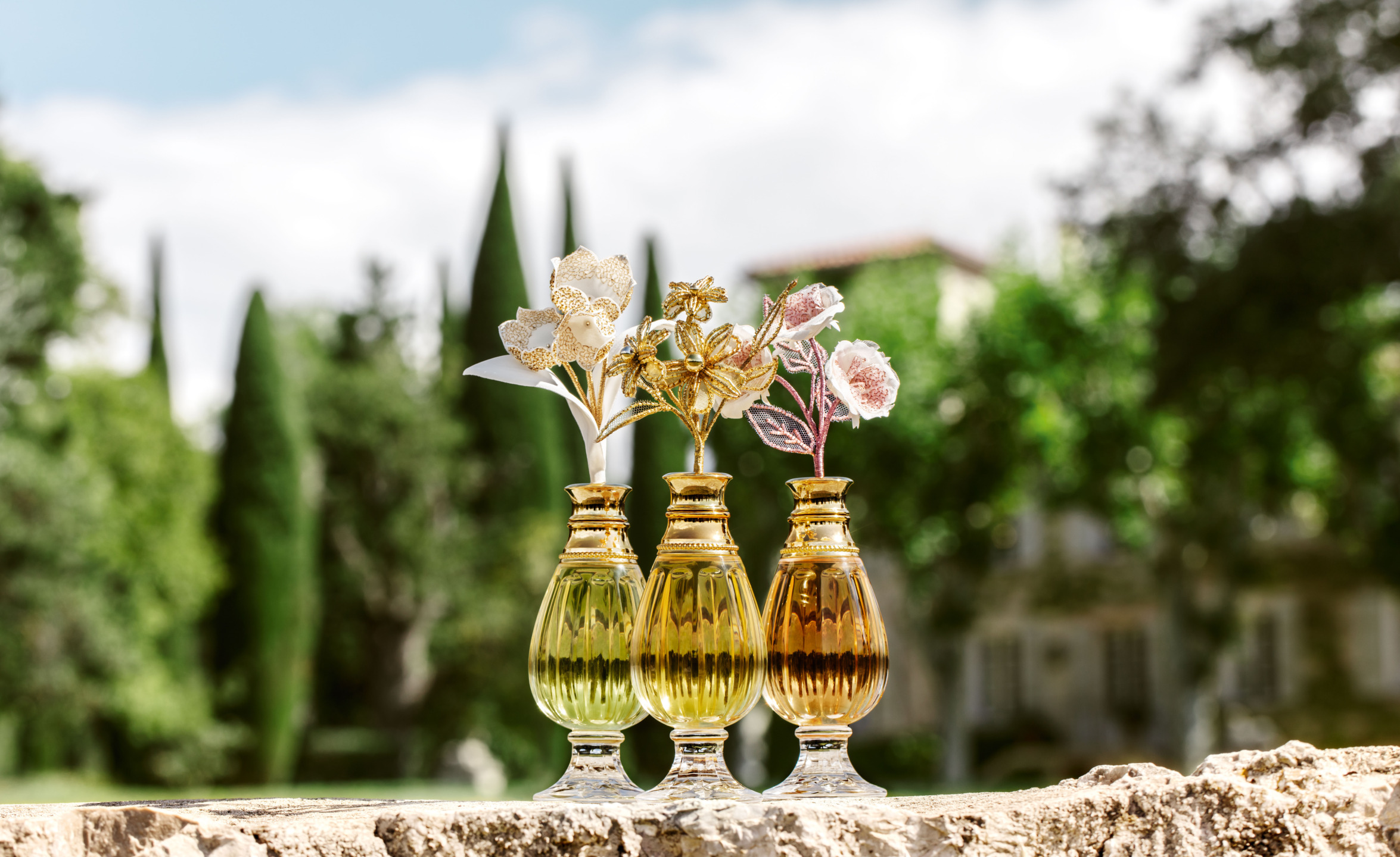 Three gold glass vases with decorative flowers on a stone ledge, green trees and blurred house in the background.