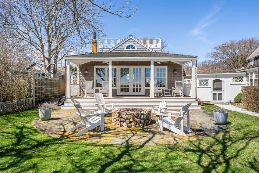 Cozy house with large porch, Adirondack chairs, and stone fire pit on a sunny day with clear blue skies.
