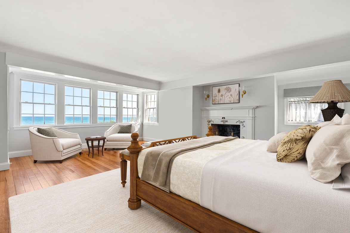 Spacious bedroom with ocean view, large windows, cozy seating, wooden bed, fireplace, and soft neutral decor.