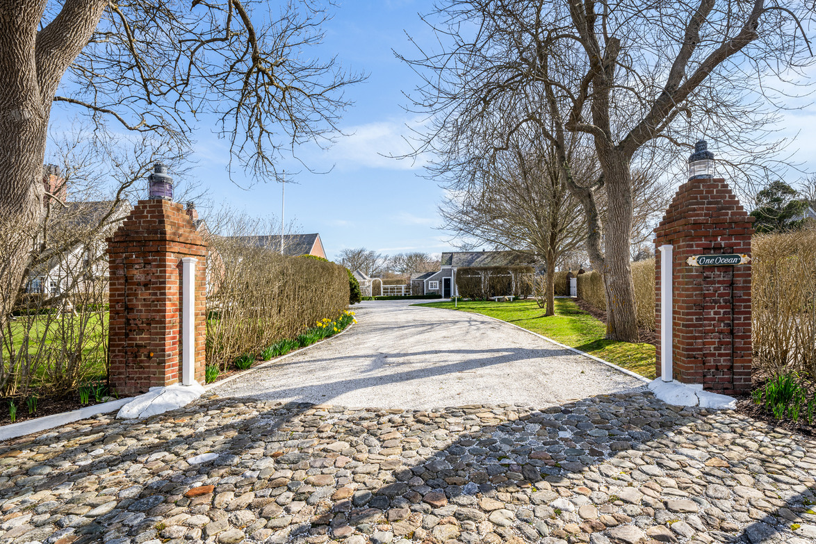Cobblestone driveway entrance with brick pillars, bare trees, and a distant house on a sunny day.