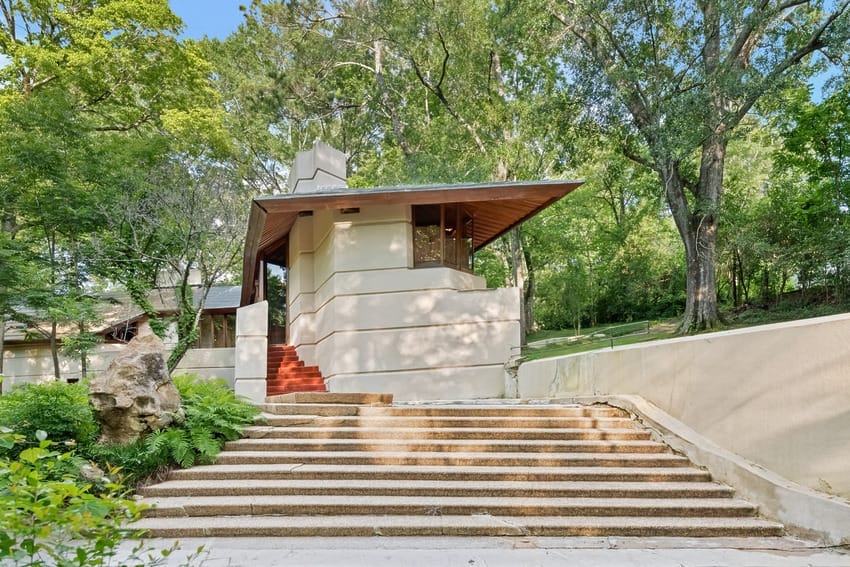 Modern, angular house with stone steps set among lush green trees, featuring a unique roof design and minimalist architecture.