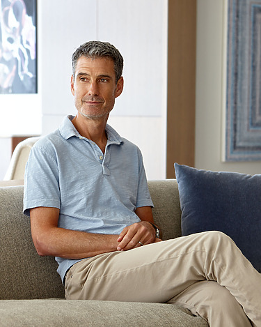 Man in a light blue polo shirt sitting on a sofa with a thoughtful expression in a modern living room setting.
