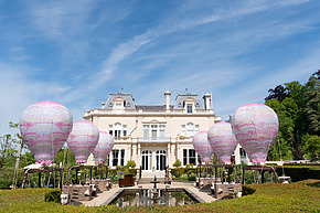 Elegant mansion with pink hot air balloon sculptures and reflecting pool in a landscaped garden under a blue sky.