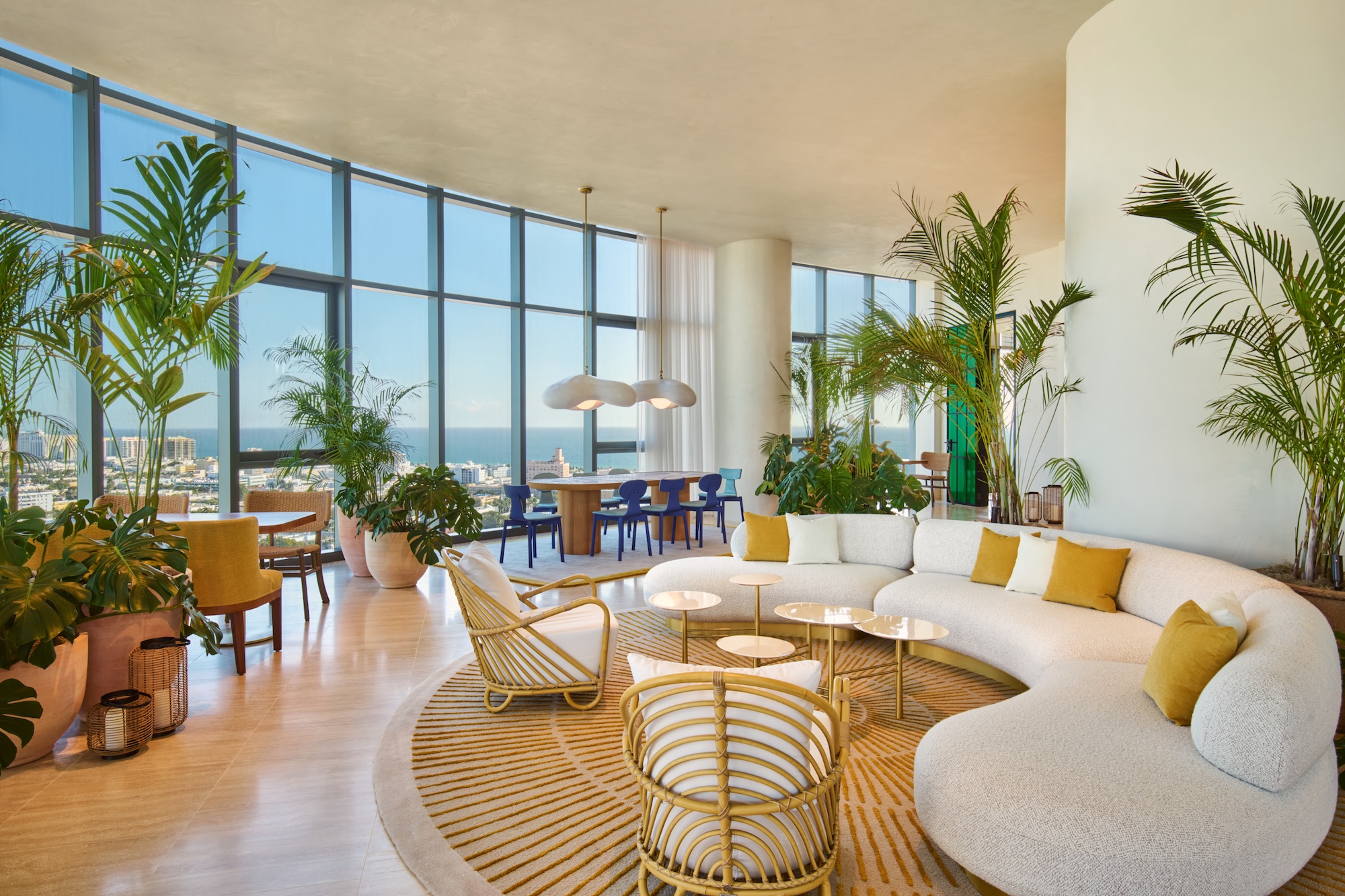 Spacious modern living room with large windows, round sofa, indoor plants, and a dining area overlooking a cityscape.