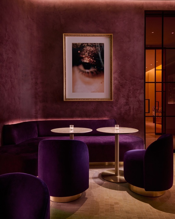 Elegant lounge with purple velvet seating, round tables, and framed eye artwork on rich burgundy walls.