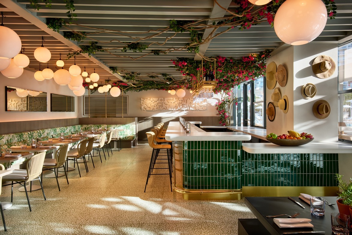Modern restaurant interior with green-tiled bar, hanging plants, and globe lighting, featuring tables and seating.