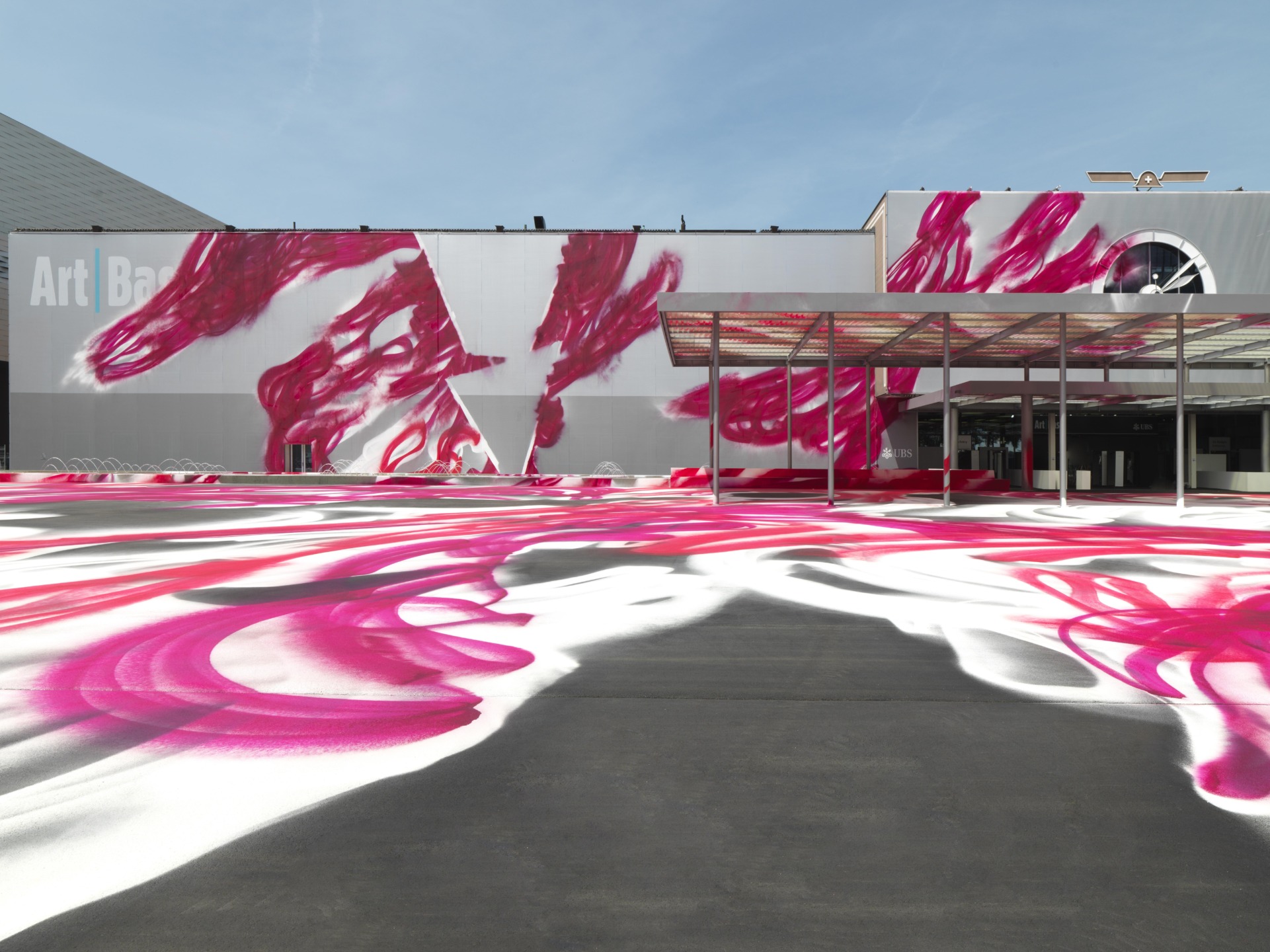 Large building facade with abstract pink and white artwork, extending across the ground, under a clear blue sky.