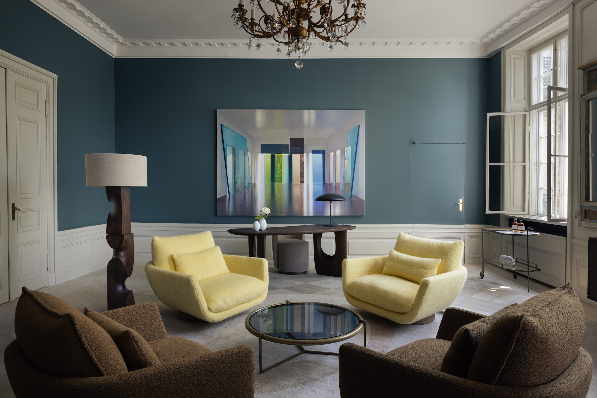 Modern living room with brown and yellow armchairs, a round glass coffee table, wall art, and large windows.