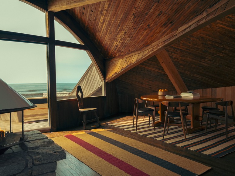 Cozy wooden room with ocean view, featuring a dining table, chairs, a stone fireplace, and colorful striped rug.