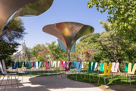 Colorful chairs arranged in a circle outdoors under large metallic structures with trees and plants in the background.