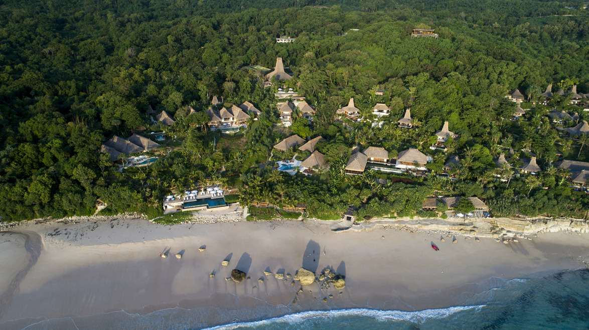 Aerial view of beachside resort with lush greenery and thatched-roof villas surrounded by dense forest, near the ocean.