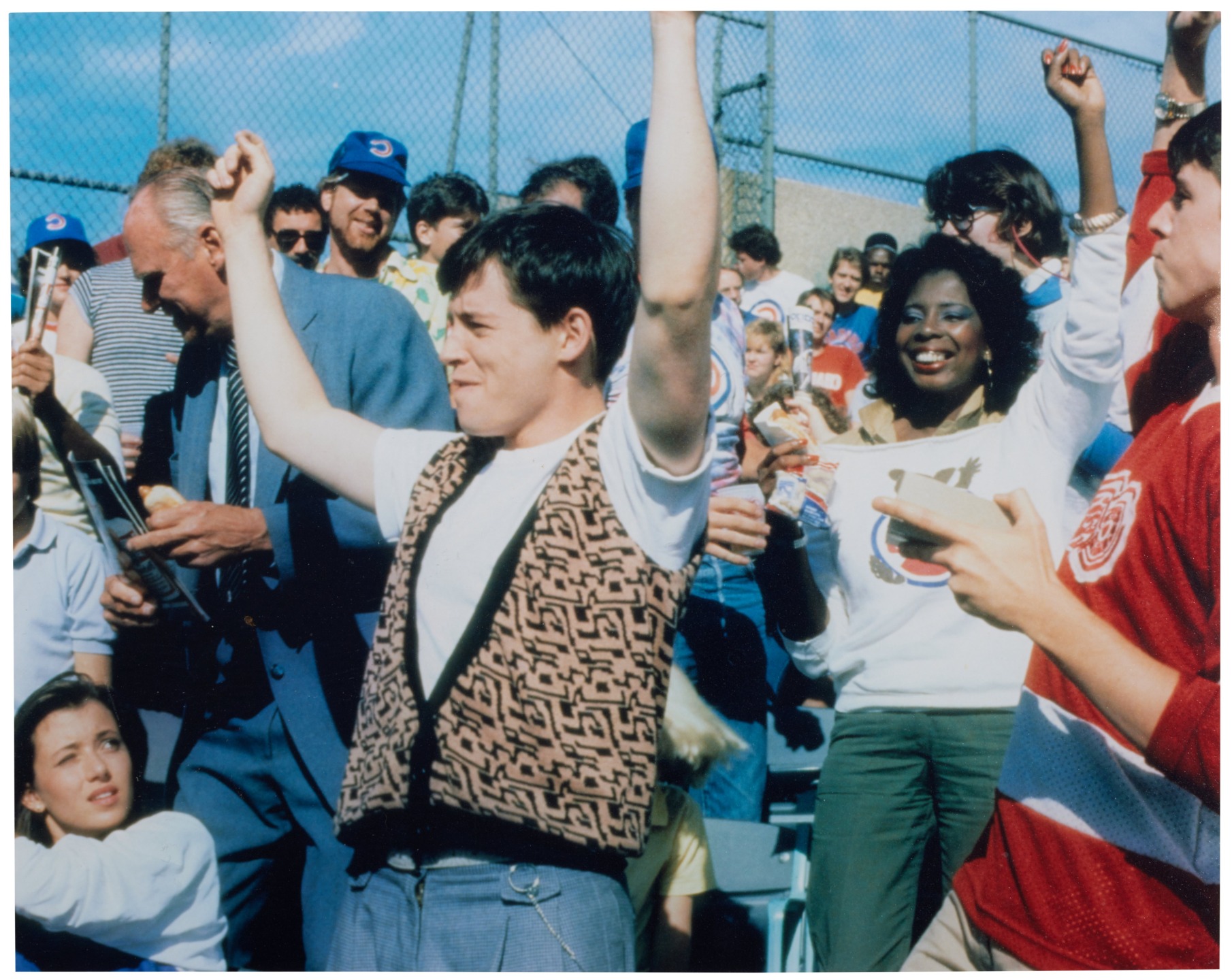 People cheering at a sports event in a stadium, wearing colorful clothing, with blue sky and a chain-link fence in the background.