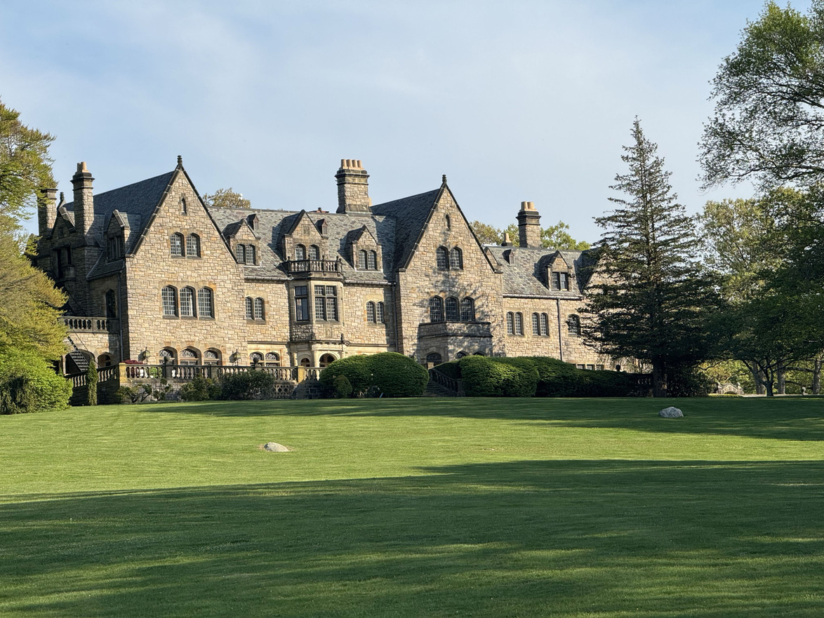 Historic stone mansion with slate roof surrounded by lush green lawn and trees on a clear day