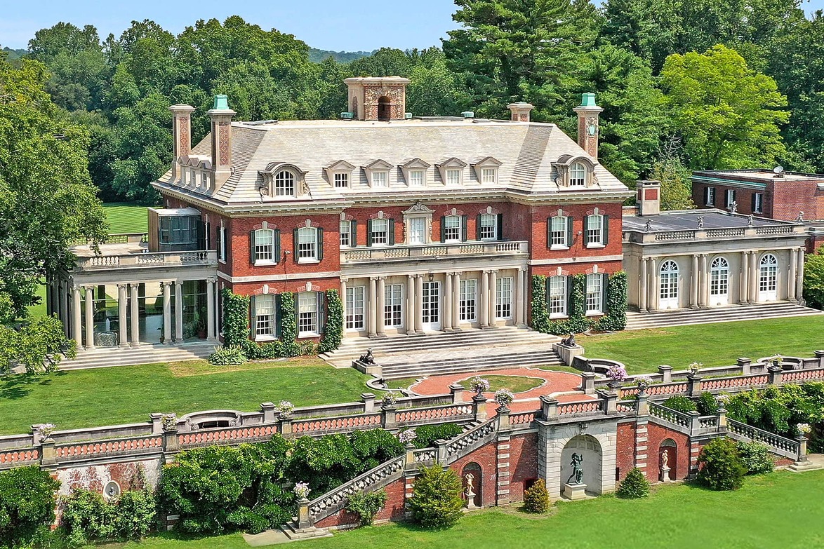 Large brick mansion with classic architecture, surrounded by lush trees and manicured gardens, on a sunny day.