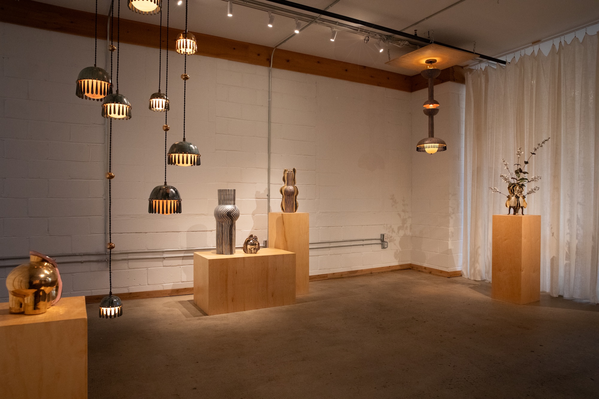 Artistic gallery space with hanging lamps and sculptures on wooden pedestals, set against a white brick wall and curtain backdrop.