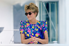 Person wearing floral dress and sunglasses with short hair stands on a balcony, smiling.