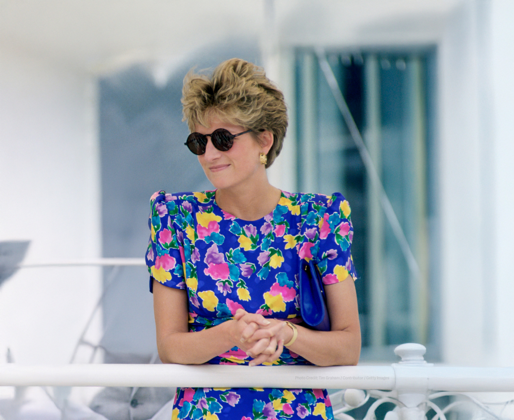 Person wearing floral dress and sunglasses with short hair stands on a balcony, smiling.