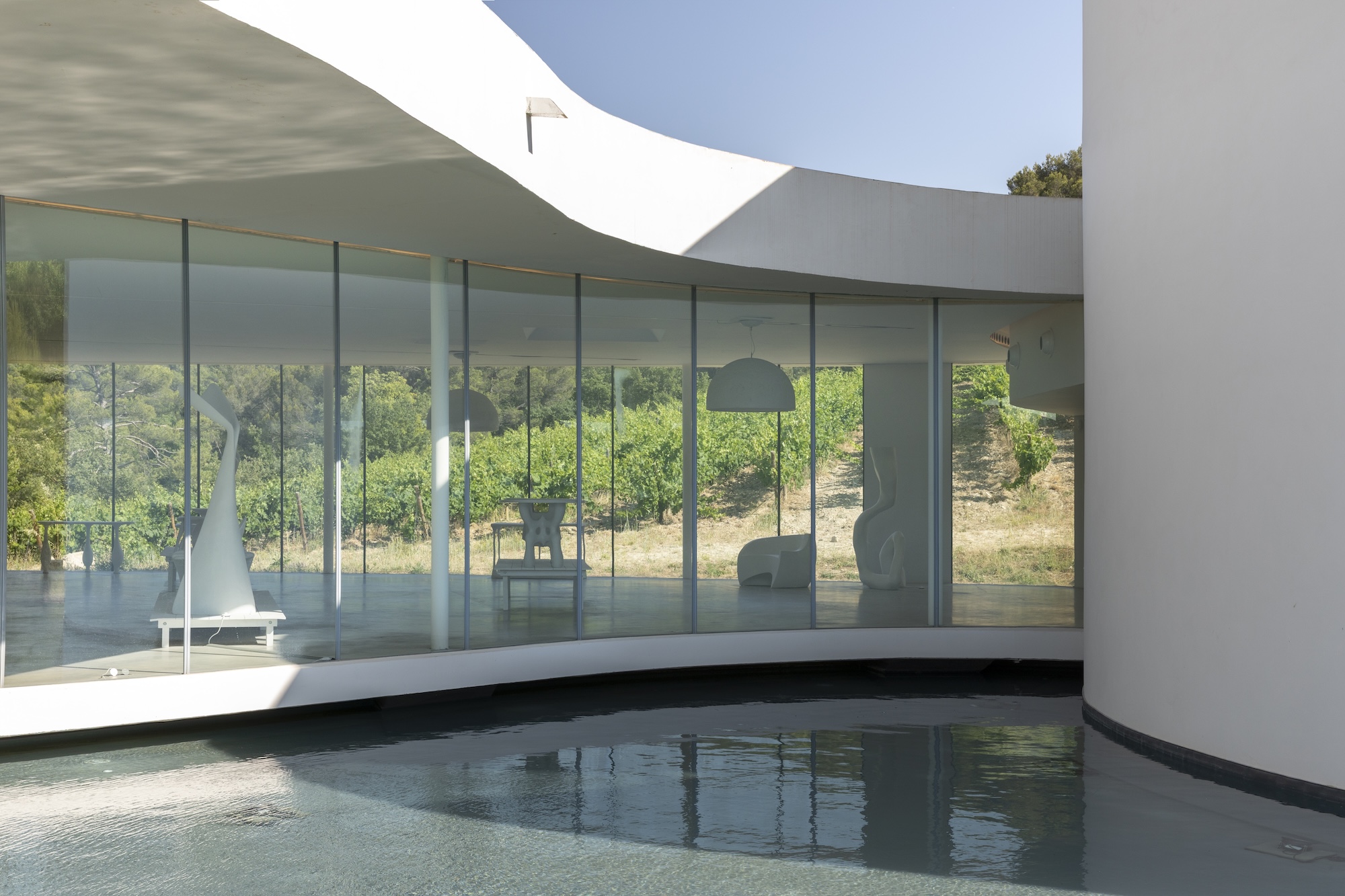 Modern house with floor-to-ceiling glass walls and curved architecture overlooking a pool and greenery outside.