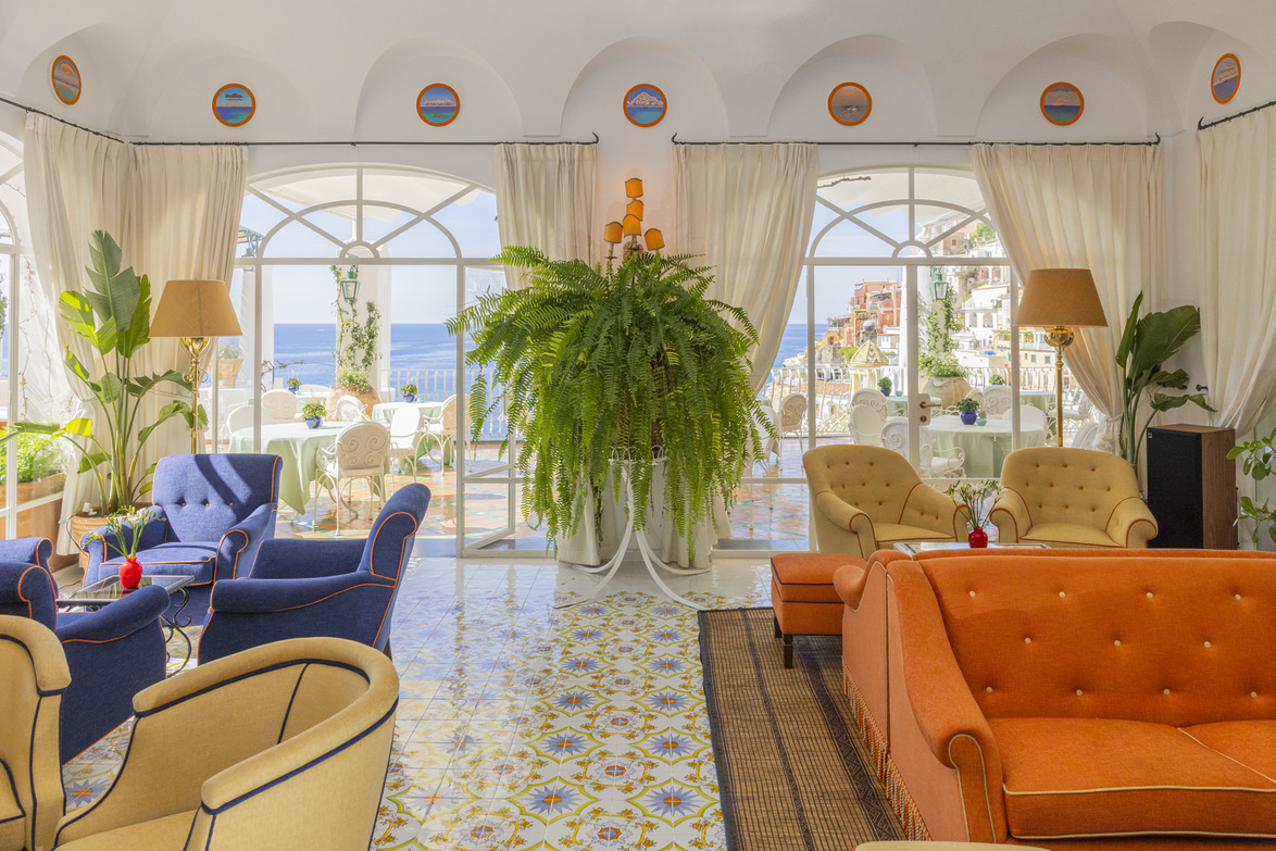 Bright, elegant lounge with colorful sofas, large windows overlooking a scenic seaside view and lush greenery.