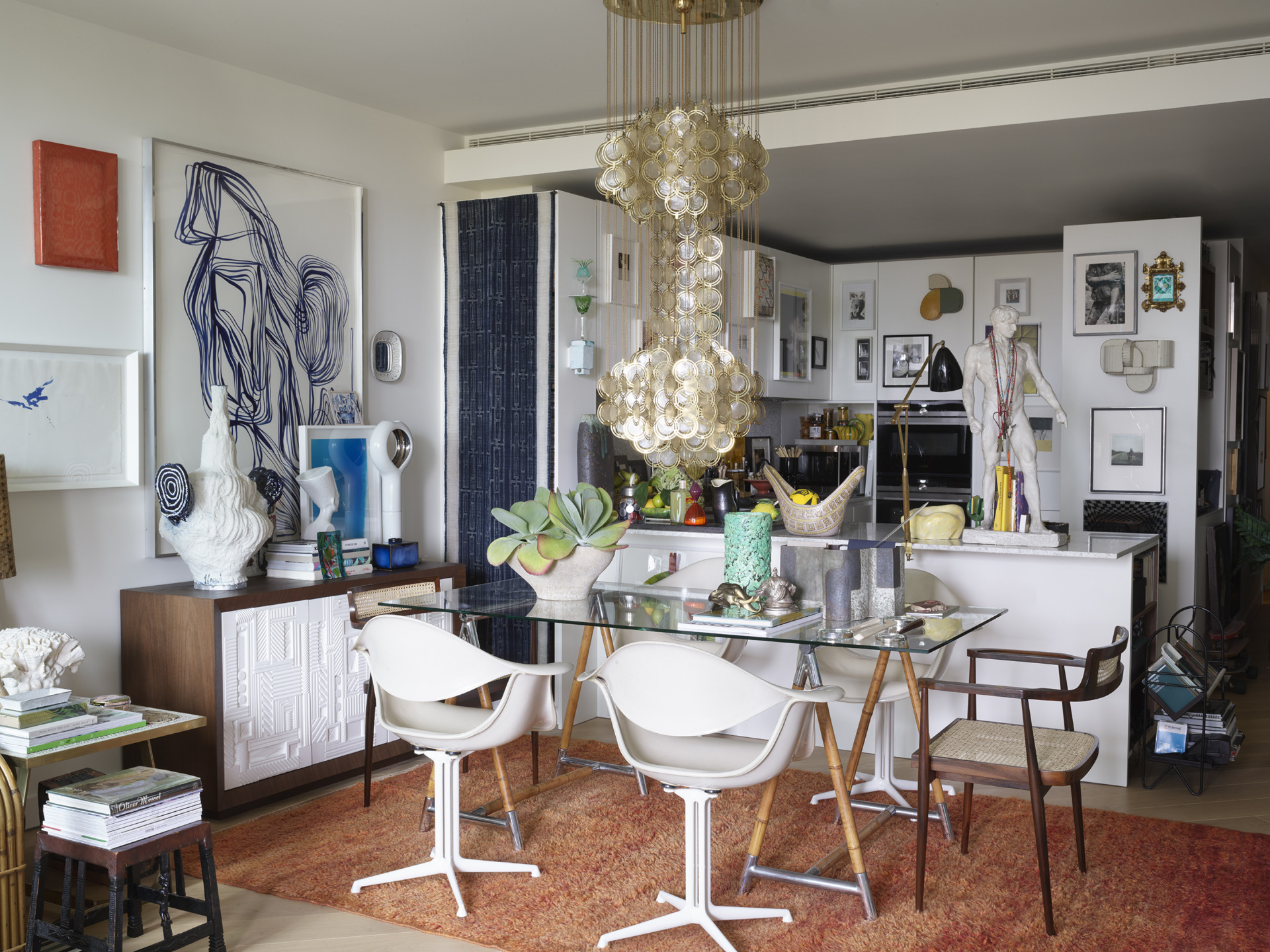 Modern dining room with eclectic decor, art on walls, glass table, white chairs, wooden elements, and a textured chandelier.