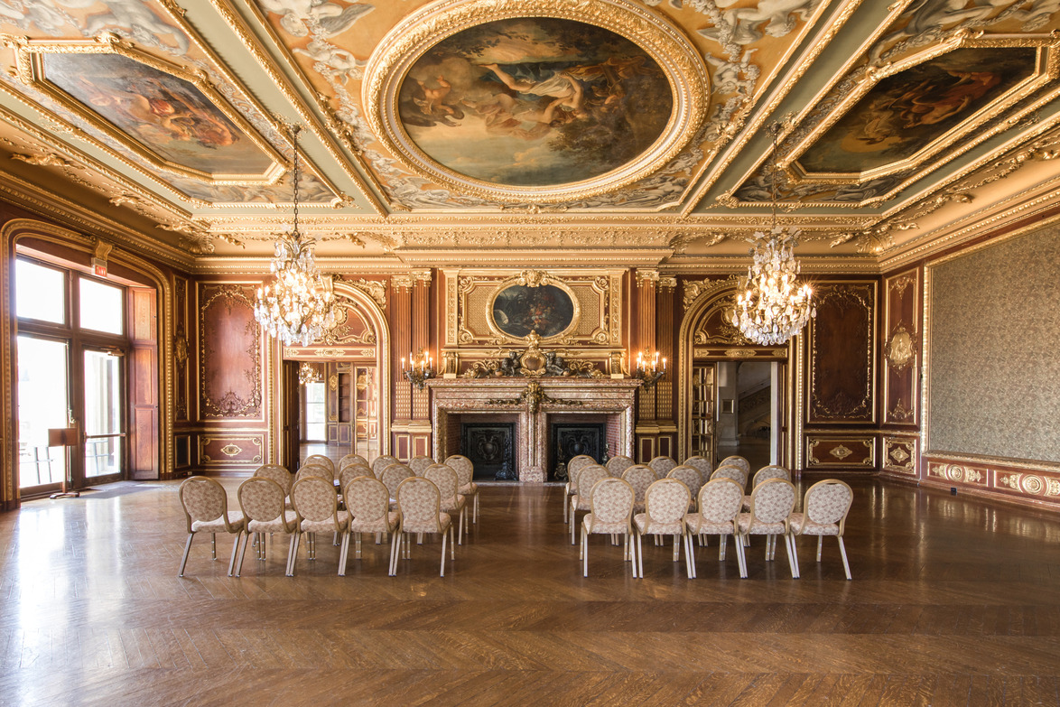 Ornate ballroom with chandeliers, intricate ceiling artwork, rows of chairs facing a decorated fireplace in an elegant setting.