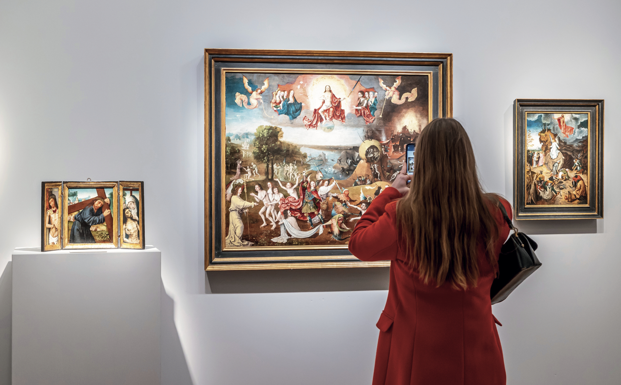 Person in a red coat viewing and photographing classical paintings in an art gallery.