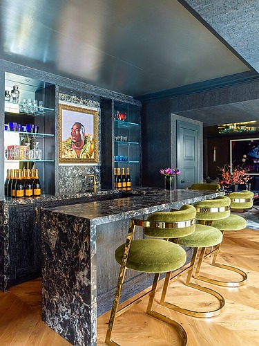 Luxury home bar with marble countertop, green bar stools, and artwork on the wall.