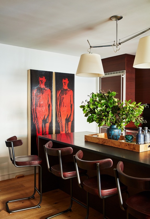 Modern kitchen with abstract art, four chairs at a dark counter, potted plants, blue vases, and overhead lighting.