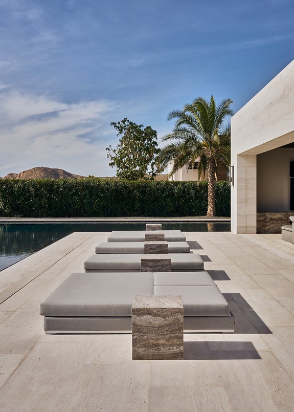 Modern outdoor patio with minimalistic lounge chairs, stone tables, and a swimming pool surrounded by greenery and palm tree.