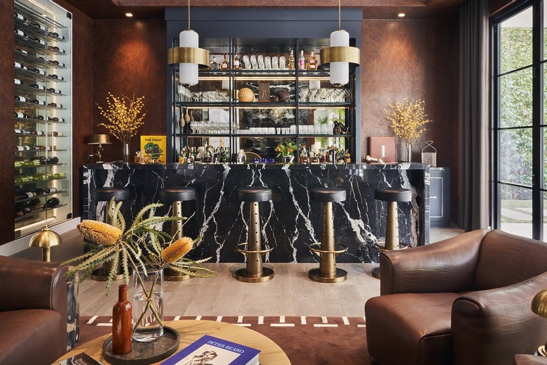 Luxury home bar with marble countertop, leather stools, wine rack, and cozy seating area with decorative plants.