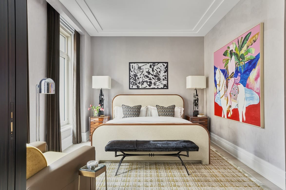 Luxurious bedroom with a stylish bed, vibrant artwork, plush armchair, and large window with curtains.