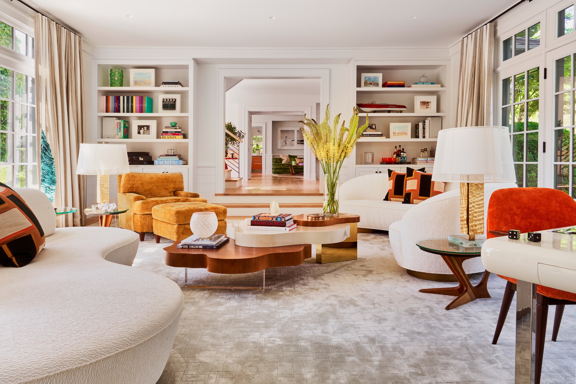 Modern living room with colorful furniture, large windows, bookshelves, and a central coffee table with a vase of yellow flowers.