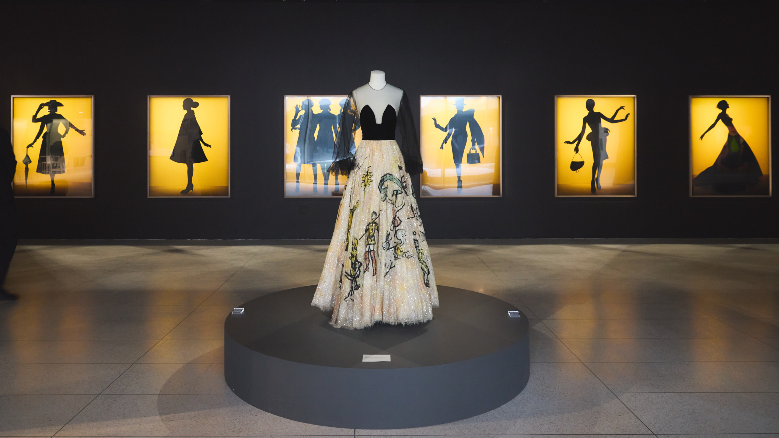 Exhibit display featuring a mannequin wearing a black and floral gown, silhouetted fashion posters in the background.