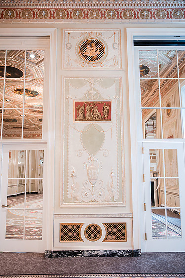 Ornate interior wall with intricate gold details and mirrored doors reflecting an elegant room.