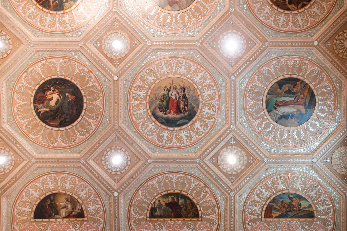 Ornate ceiling with patterned tiles and round paintings depicting historical religious scenes, illuminated by soft lighting.