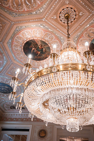 Elegant crystal chandelier with ornate ceiling and detailed paintings in a luxurious room.
