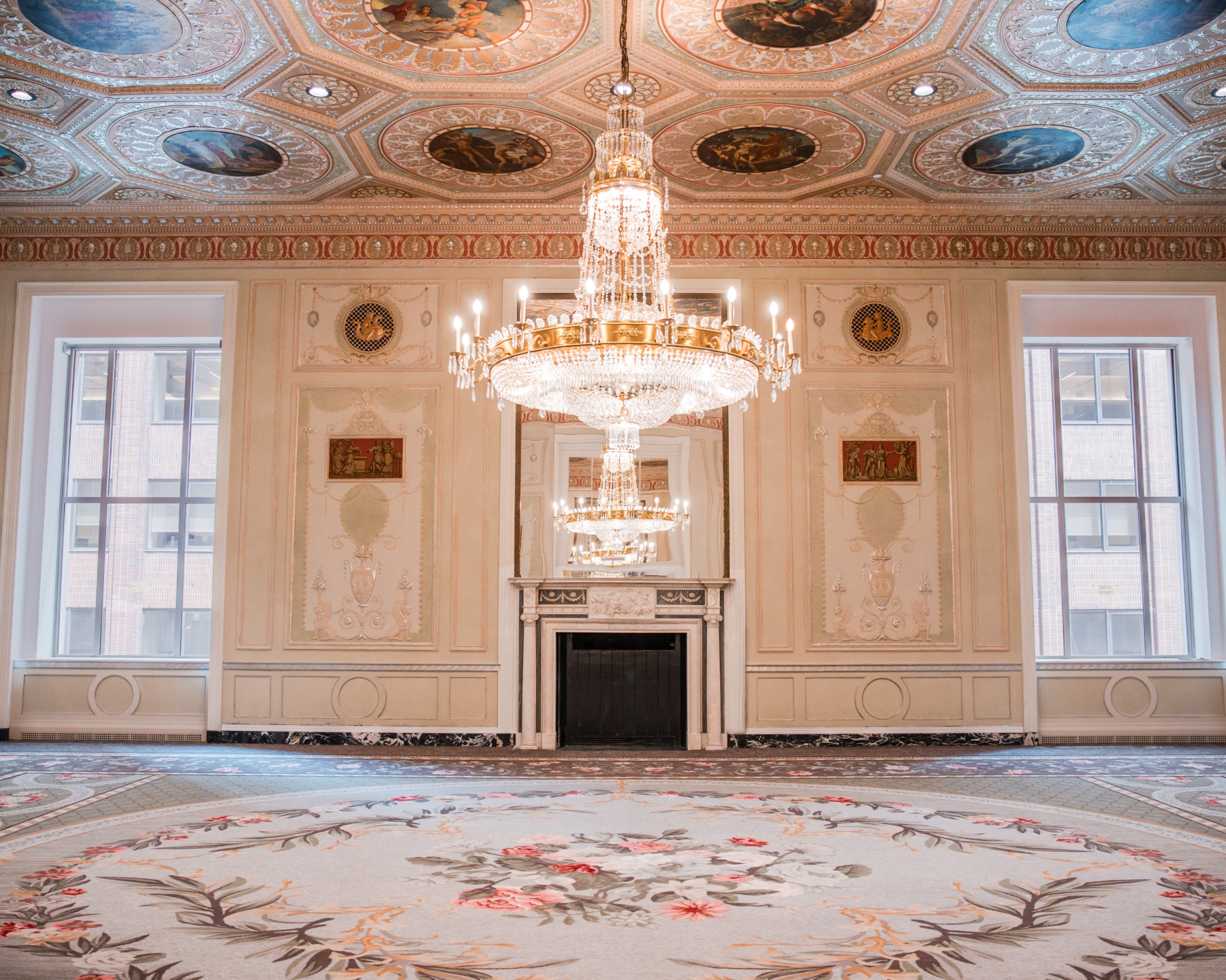 Elegant ballroom with ornate chandeliers, decorative ceiling, and intricate flooring, featuring large windows and a fireplace.