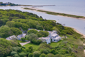 Aerial view of a large coastal house surrounded by greenery and water near a beach.