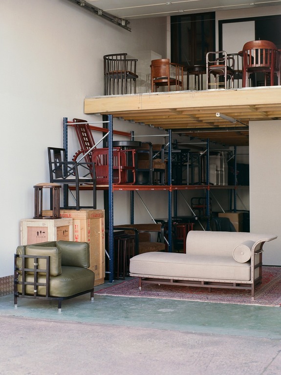 Warehouse interior with vintage chairs on metal shelves, a green armchair, and a beige chaise longue on the floor.