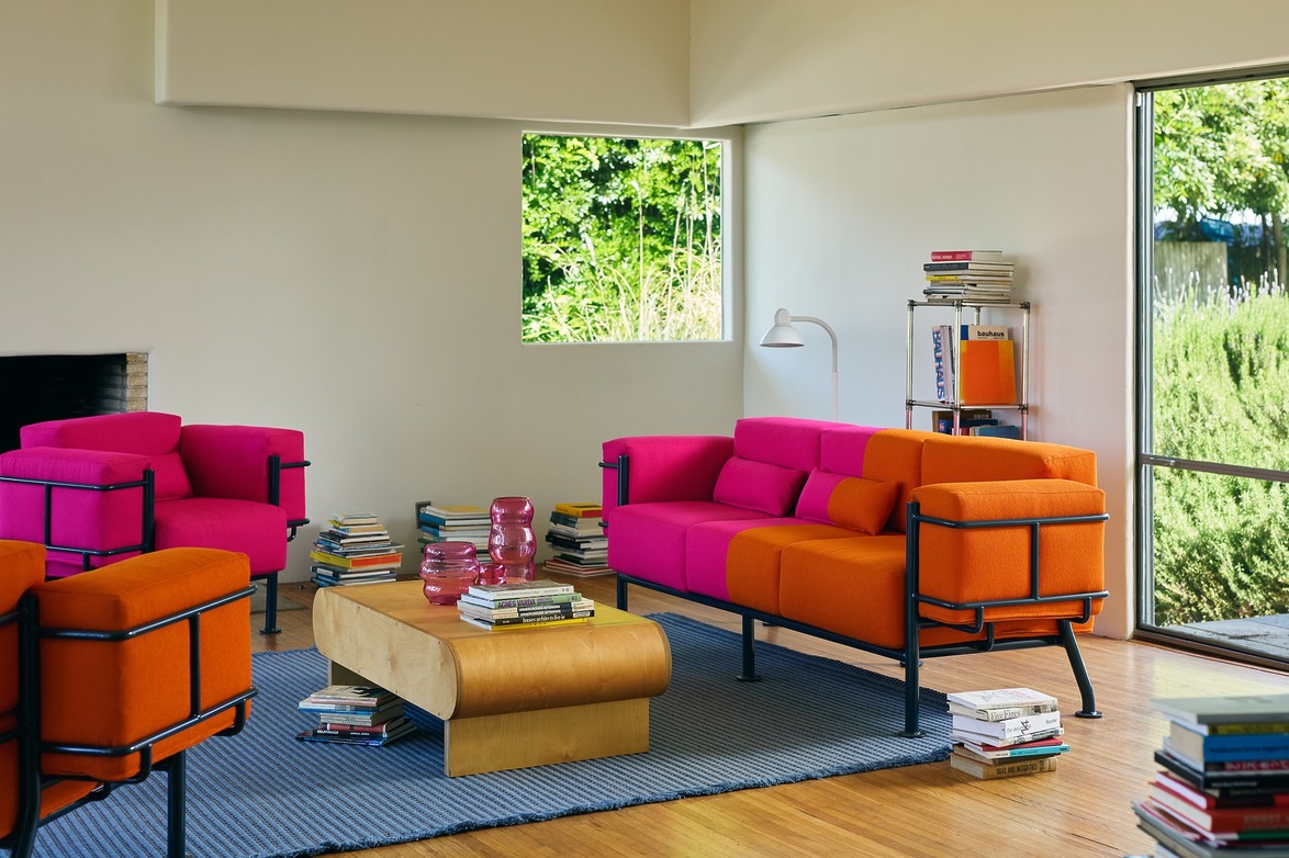 Modern living room with bold pink and orange sofas, wooden coffee table, books stacked, and a window with garden view.