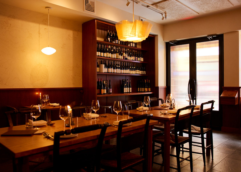 Cozy restaurant interior with elegant dining table, chairs, wine shelves, soft lighting, and large windows.