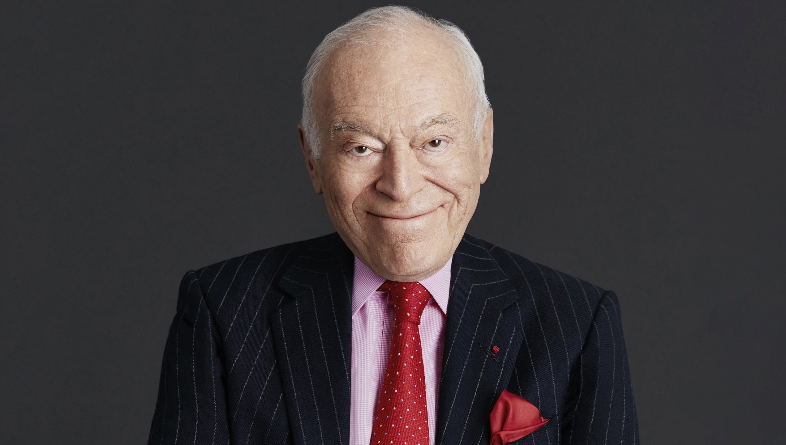 Elderly man in a striped suit and red tie, smiling against a dark background.
