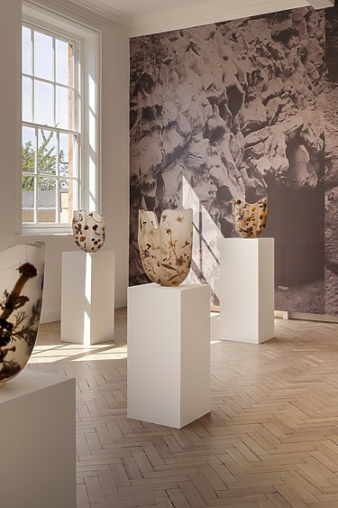 Art gallery installation with floral-themed vases on pedestals and a black and white nature mural in the background.