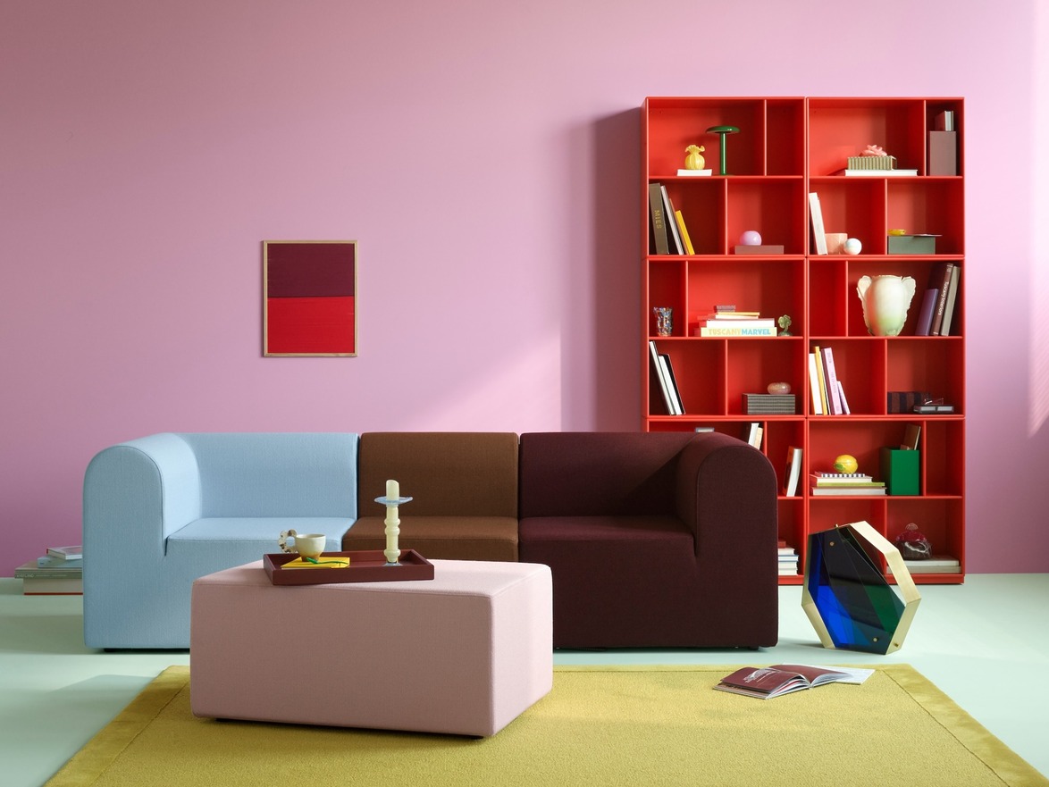 Modern living room with blue and brown sofa, pink ottoman, colorful bookshelf, and abstract art on a pink wall.