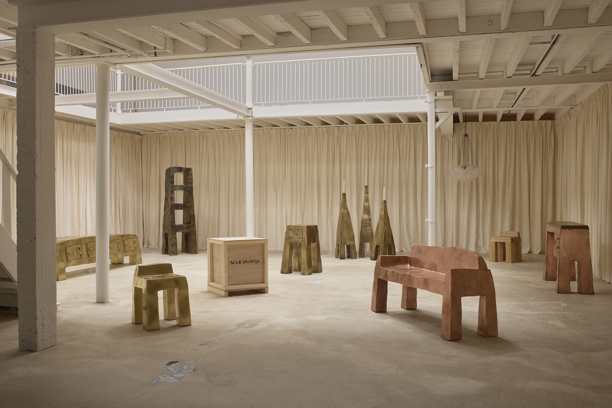 A spacious gallery with wooden stools, benches, and sculptures, surrounded by beige curtains and white beams.