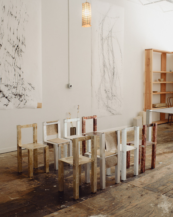 Minimalist wooden chairs arranged in a rustic art studio with abstract wall art and a wooden bookshelf.