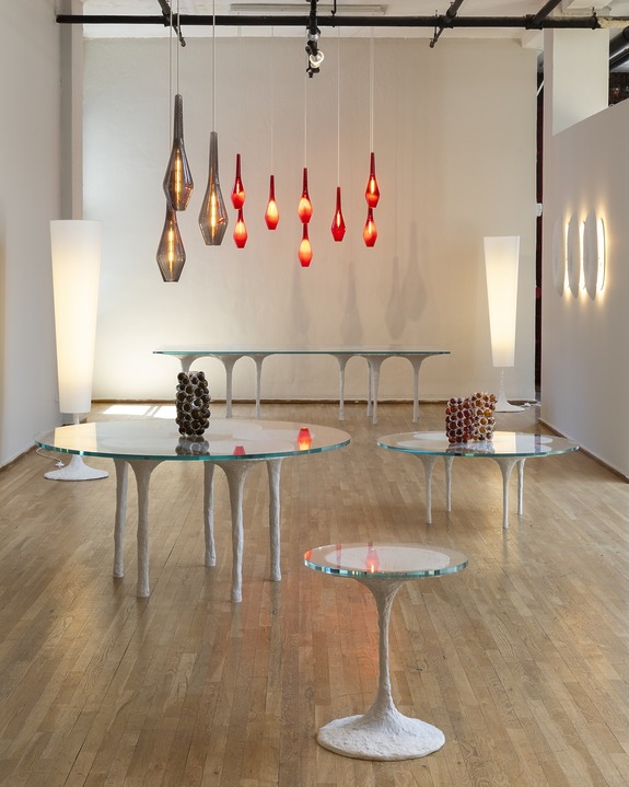 A gallery display featuring modern glass-top tables and vibrant red pendant lights in a minimally decorated room.