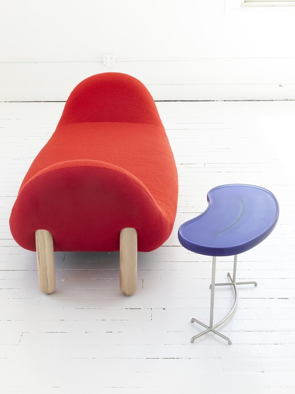 Red chaise lounge with wooden legs next to a small, purple kidney-shaped table on a white wooden floor.