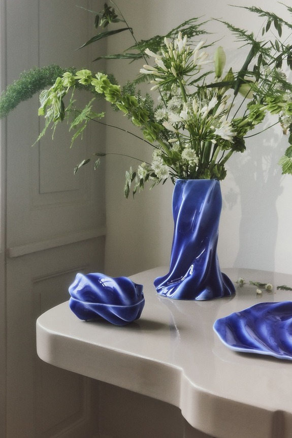 Blue ceramic vase with green and white flowers on a white table, next to matching blue ceramic pieces.