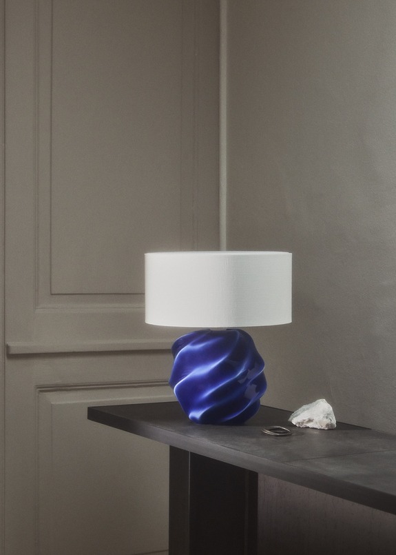 Modern table lamp with blue sculpted base and white lampshade on dark wooden table beside a small rock in a minimalist room.